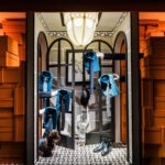 Hermès Vitrines d'Automne 2024 Lever de rideaux - room entirely covered in orange Hermès boxes with jeans shirt and tie on coat rack, boots and dog at centre